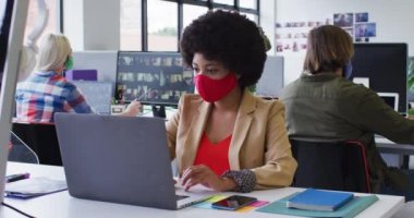 Modern ofiste dizüstü bilgisayar kullanarak maske takan melez bir iş kadını. Arka plandaki iş arkadaşları. İş yerindeki hijyen, kovid 19 koronavirüs salgını sırasında.