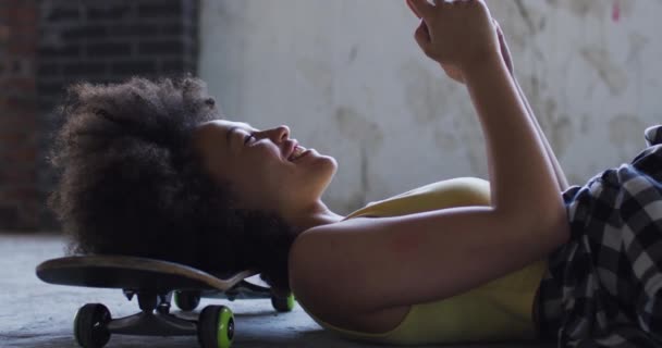 Femme de race mixte dans un bâtiment vide couché sur une planche à roulettes en riant smartphone — Video