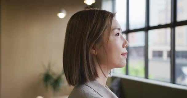 Asiática mujer de negocios de pie mirando por la ventana en la oficina moderna — Vídeos de Stock
