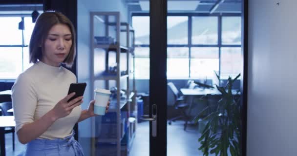 Asiatische Geschäftsfrau läuft mit Smartphone und Kaffeetasse im modernen Büro — Stockvideo