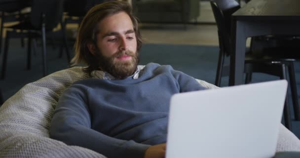 Hombre de negocios caucásico sentado en el puf con un ordenador portátil en la oficina moderna — Vídeo de stock