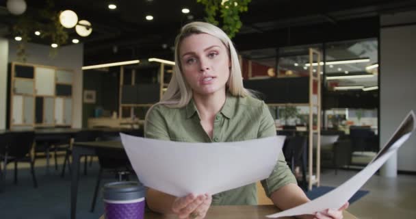 Mulher de negócios caucasiana tendo um bate-papo por vídeo passando pela papelada no escritório moderno — Vídeo de Stock