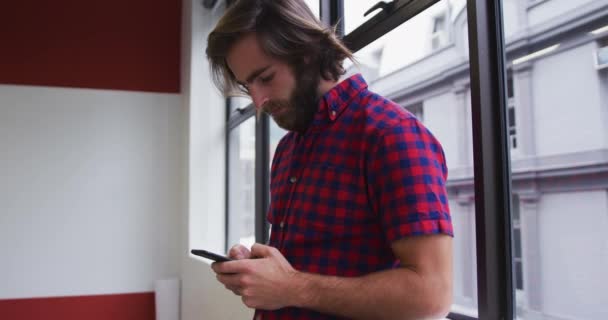 Hombre de negocios caucásico de pie con teléfono inteligente mirando por la ventana en la oficina moderna — Vídeos de Stock