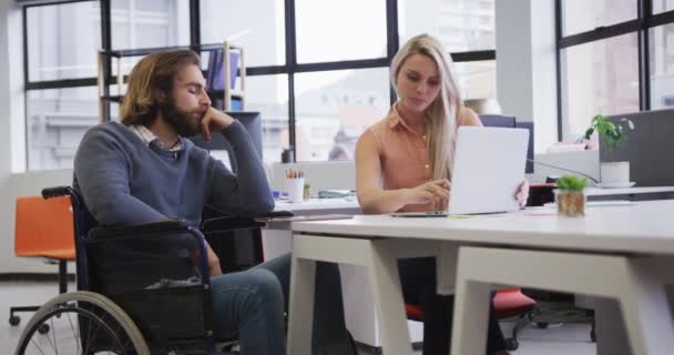 Hombre de negocios caucásico discapacitado sentado en silla de ruedas usando el ordenador portátil hablando con su compañero de trabajo en la oficina — Vídeos de Stock