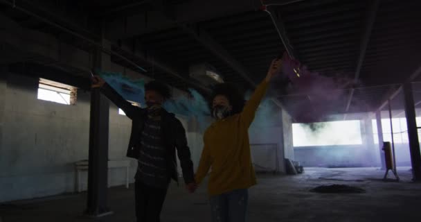 Homme et femme de race mixte tenant des fusées éclairantes bleues et violettes marchant dans un bâtiment vide — Video