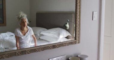 Beyaz kadın yüzüne dokunuyor. Aynaya bakıyor. Evdeki yatakta oturuyor.