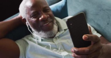 Afro-Amerikalı adam evde kanepede yatarken akıllı telefon kullanıyor.