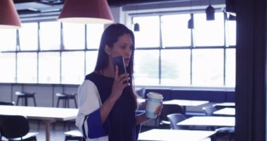 Karışık ırk işkadını akıllı telefondan konuşuyor elinde kahve, ofis koridorunda yürüyor. Modern ofiste çalışan iş adamları.