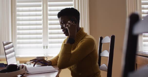 Mujer afroamericana hablando en smartphone y usando laptop mientras trabaja desde casa — Vídeos de Stock