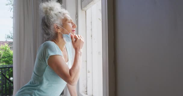 Starší běloška se spuštěnou obličejovou maskou, dívající se z okna domů — Stock video