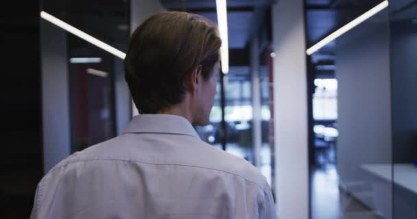 Vista trasera del hombre de negocios caucásico caminando por el pasillo de la oficina sosteniendo el teléfono inteligente — Vídeo de stock