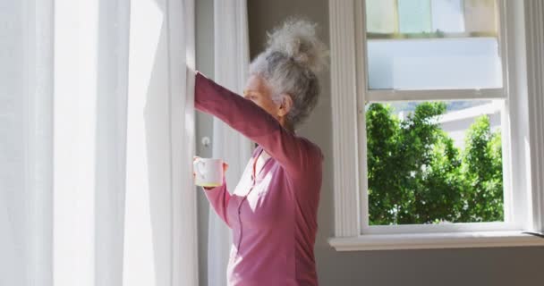 Старшая белая женщина с кофейной чашкой, открывающими занавески на окне дома — стоковое видео