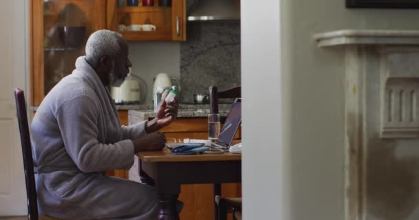 Homme âgé afro-américain tenant des médicaments vides tout en ayant un appel vidéo sur ordinateur portable à la maison — Video