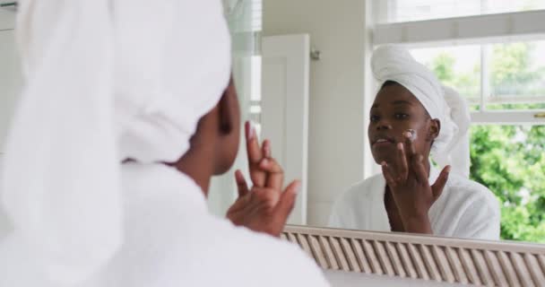 African american woman in bathrobe applying face cream while looking in the mirror at home — Stock Video