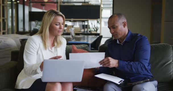 Dos colegas de negocios diversos usando el ordenador portátil y discutiendo el papeleo en una reunión informal — Vídeos de Stock