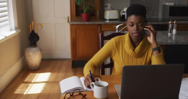 Afrikai amerikai nő beszél okostelefonon, és jegyzetel, miközben otthon dolgozik — Stock videók