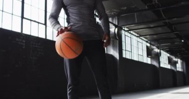 Afrikalı Amerikalı bir adam boş bir binada durup basketbol topuyla oynuyor ve kameraya bakıyor.