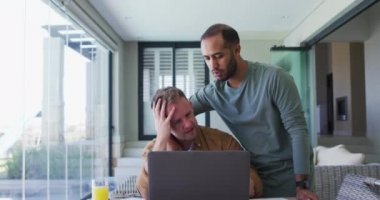 Çok ırklı erkek, aynı cinsiyetten çift evde oturmuş dizüstü bilgisayar kullanıyor ve kucaklaşıyorlar.