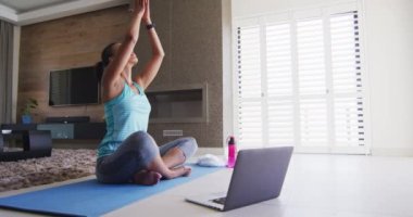 Karışık ırktan bir kadın laptopla paspasla antrenman yapıyor. Evde kaliteli vakit geçirmenin keyfini çıkarıyor. Karantina tecridinde..