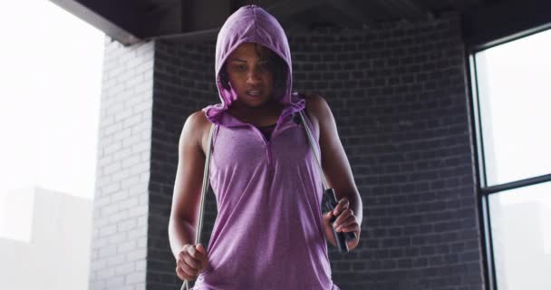 Portrait of african american woman holding skipping rope wearing hoodie looking at camera in an empt — Stok Video