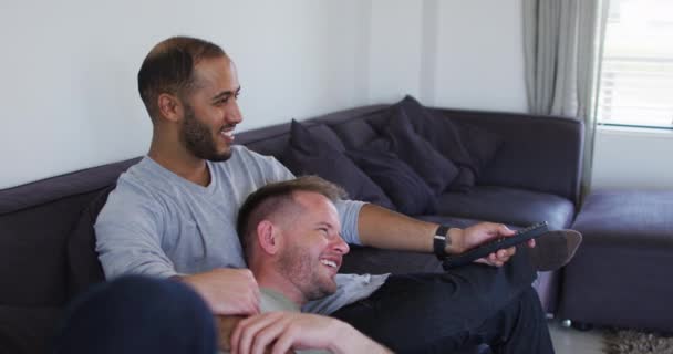 Мультинациональная гей-пара сидит на диване и смотрит телевизор — стоковое видео