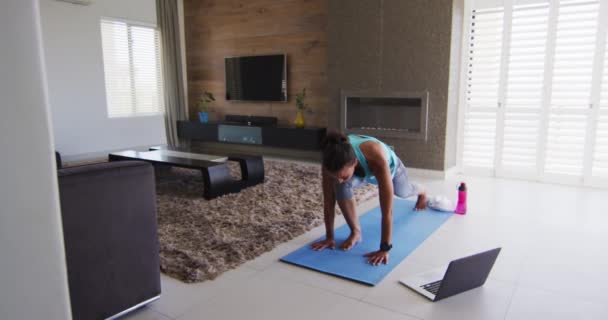 Mujer de raza mixta haciendo ejercicio en una alfombra con portátil — Vídeos de Stock