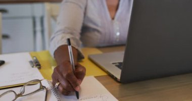 Kıdemli Afro-Amerikan kadın telefon kulaklığı takıyor. Evde dizüstü bilgisayar kullanırken not alıyor.