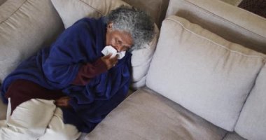 Hasta kıdemli Afrikalı Amerikalı kadın. Battaniyeyle kaplanmış. Evdeki kanepede yatarken hapşırıyor.