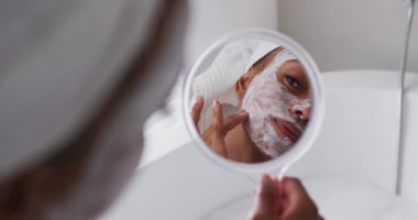 Afro-Amerikalı bir kadın banyoda ayna tutarken yüz maskesi takıyor.