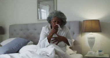 Hasta kıdemli Afrikalı Amerikalı kadın evde yatakta otururken öksürüyor.