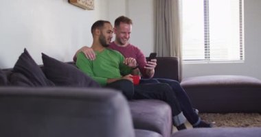 Çok ırklı eşcinsel çift kanepede oturup akıllı telefonlara bakıyorlar.