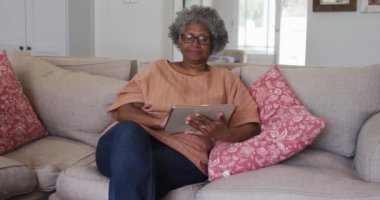 Kıdemli Afrikalı Amerikalı kadın evdeki kanepede otururken dijital tablet kullanıyor.