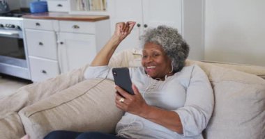 Kıdemli Afro-Amerikan kadın kulaklık takıyor. Evde akıllı telefondan video görüşmesi yapıyor.