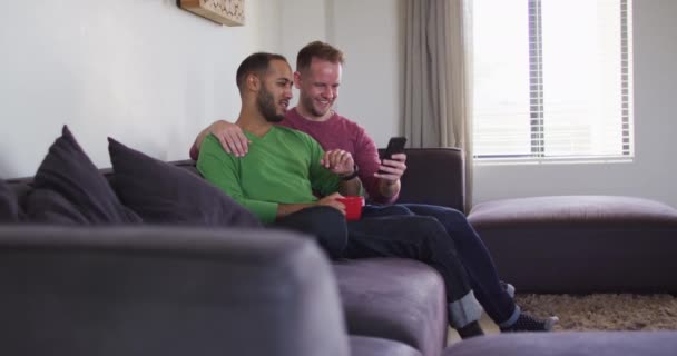 Multi etniska homosexuella manliga par sitter på soffan tittar på smartphone tillsammans — Stockvideo