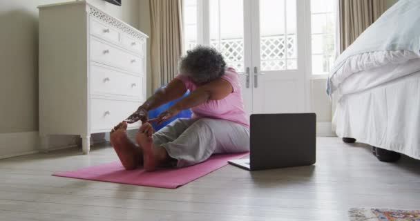 Mujer afroamericana mayor realizando ejercicio de estiramiento mientras mira el portátil en casa — Vídeos de Stock