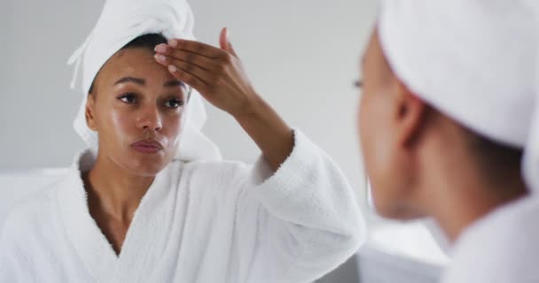 Afričanky americká žena v županu použití krém na obličej při pohledu do zrcadla v koupelně — Stock video