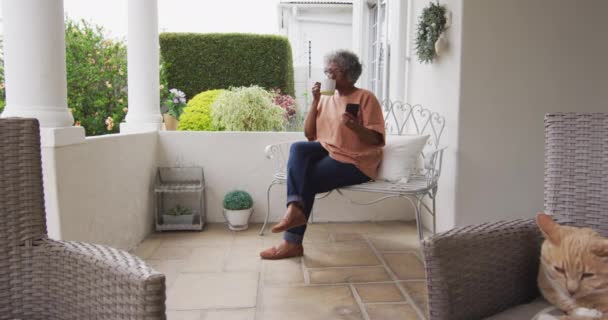 Senior αφροαμερικανή γυναίκα χρησιμοποιώντας smartphone, ενώ πίνοντας καφέ κάθεται στη βεράντα του Hou — Αρχείο Βίντεο