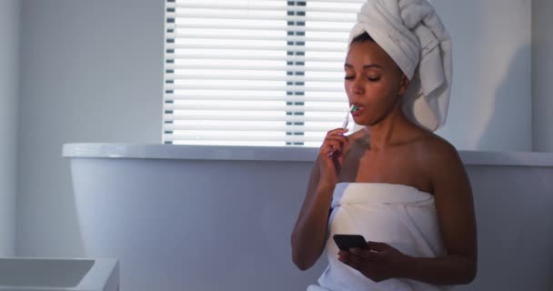 Afričanky americká žena pomocí smartphonu při čištění zubů v koupelně — Stock video