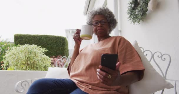 Senior africká americká žena pomocí smartphone při pití kávy sedí na verandě v hou — Stock video