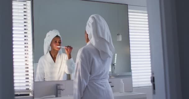 Afroamericano donna in accappatoio lavarsi i denti guardando nello specchio in bagno — Video Stock