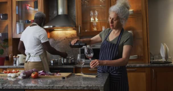 Mulher idosa caucasiana vestindo avental derramando vinho no copo na cozinha em casa — Vídeo de Stock