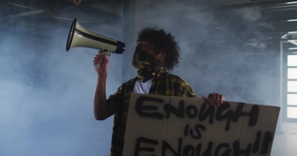 Yüz maskesi takmış Afro-Amerikan bir adam megafonla konuşuyor boş bir park yerinde protesto pankartı tutuyor. — Stok video