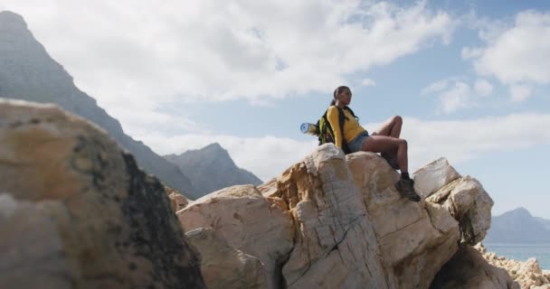 Afroamerikanerin mit Rucksack genießt beim Wandern die Aussicht sitzend auf den Felsen — Stockvideo