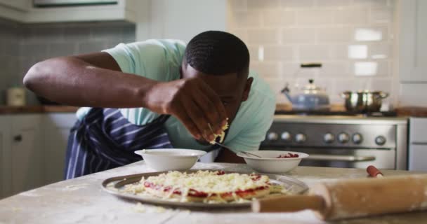 Hombre afroamericano haciendo pizza en cocina rociando queso rallado — Vídeos de Stock