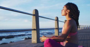 Afrika kökenli Amerikalı kadın deniz kenarındaki gezinti alanında yoga yapıyor.