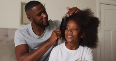 Afrikalı Amerikalı baba evde yatağında otururken kızının saçını tarıyor.