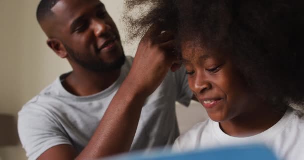 Afro-americano pai amarrando suas filhas cabelo enquanto sentado na cama em casa — Vídeo de Stock