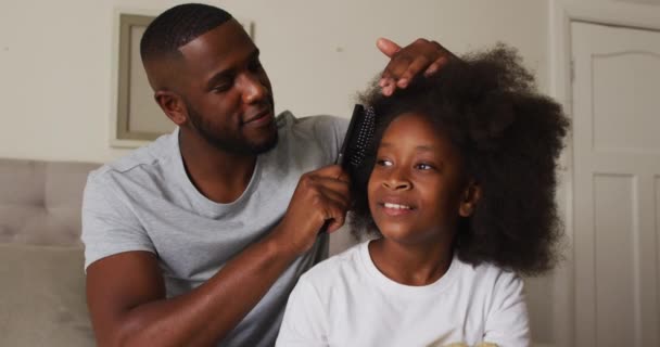 African american father brushing his daughters hair while sitting on the bed at home — Stock Video