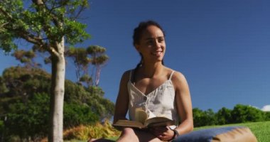 Afrikalı Amerikalı kadın çimenlerde oturmuş parkta kitap okuyor.