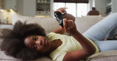 Afrikalı Amerikalı kız, arka planda babasıyla kanepeye uzanmış video oyunu oynuyor. Karantina süresince evde tek başına kalarak eğleniyor..